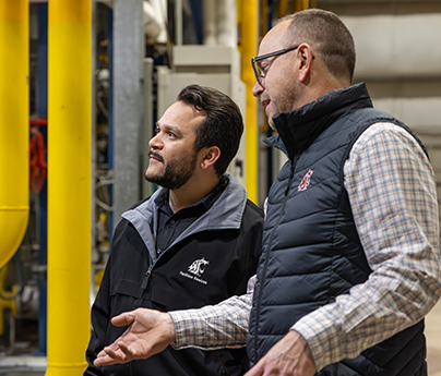 Two men wearing WSU vests are standing and chatting next to some indoor pipes.